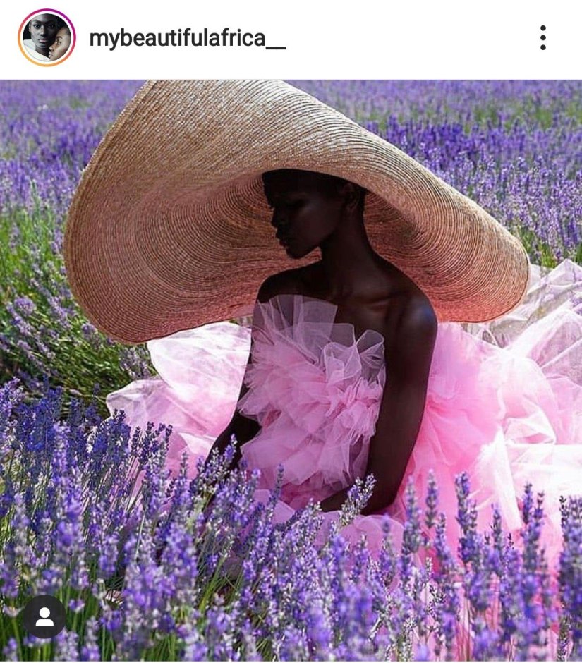 A dark skin model sits in a field of flowering Lavender. They're wearing an oversized, floopy woven hat, and a pink strapless dress made of tulle. They're eyes are turned downword.