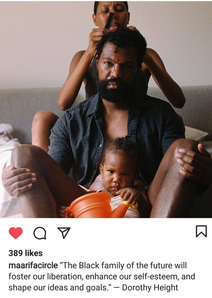 An Afro sits on the couch with their Afro lover between their legs as they style their hair. The Afro baby sits in the foreground playing with an organge pail.