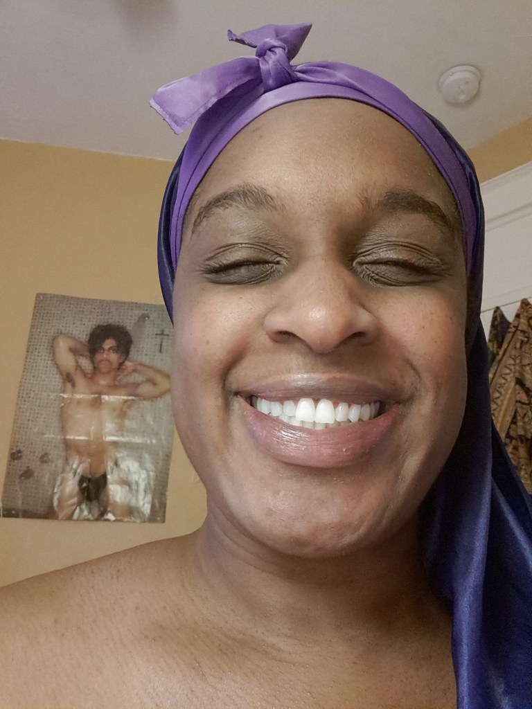 Janeé smiles with squined eyes and purple head wrap in from of a Prince poster.