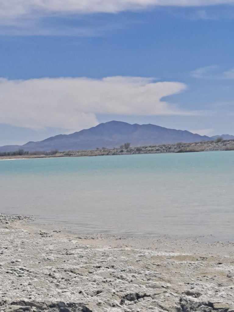 loss dirt and sand edges a periwinkle lake. In the background is a hill, whose base is lined by trees. Large white clouds in the blue sky.