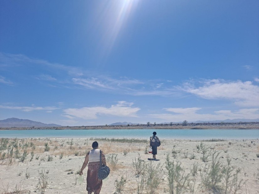2 people walk across the sand to the lakes edge. A mountain is on the horizon.