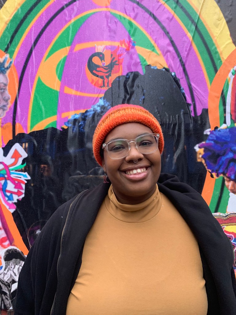 Syon (they/she/he) is an orange sweather and black jacket poses in front of a artfully painted wall