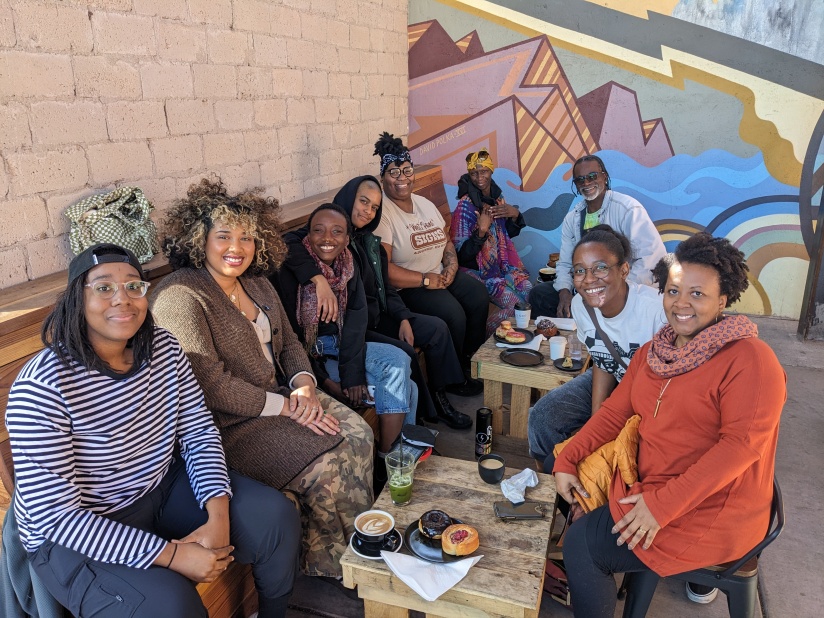 9 Afro folks mainly with long sleeve outfits, sit against a wall mural.