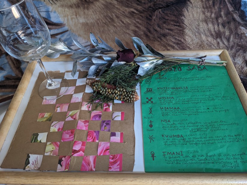 The Nguzo Saba (Seven Principles) in red on green paper. An image of pink roses in woven into cardboard to make a Mkeka (Mat). Upon which is a clear wine glass with long stem. A green and yellow Mindi (Corn Cob) is tucked into the Cedar which is in a bouquet of silver leaves and red roses.