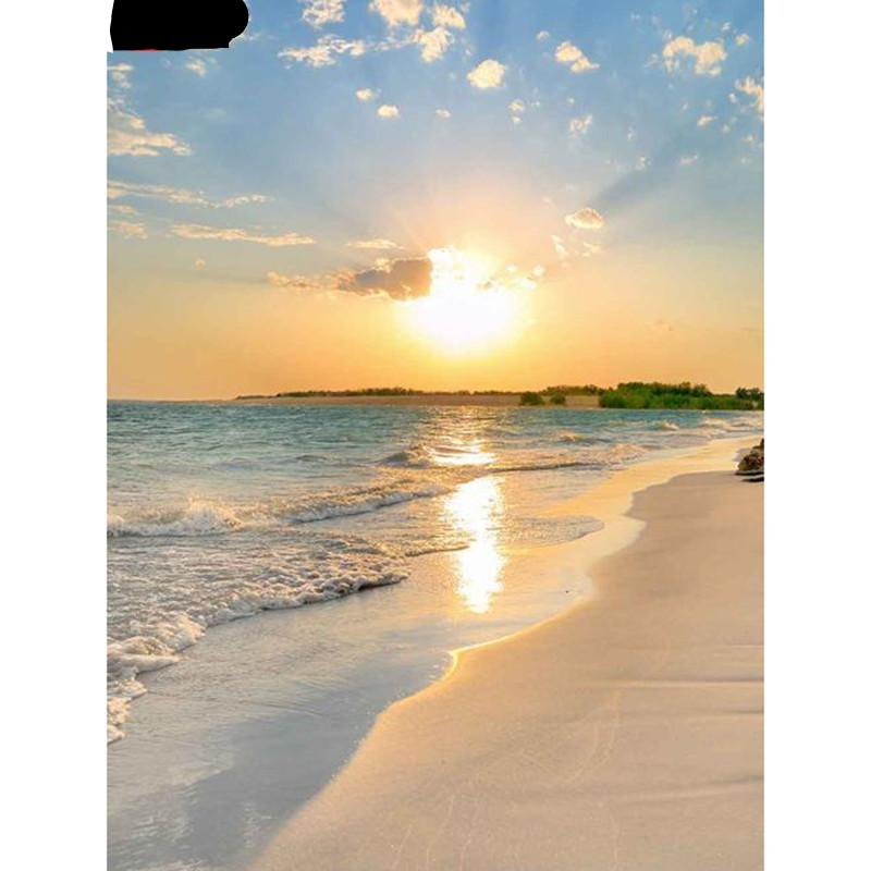 The bright, shining Sun is lowering beyond a sandy beach. The tan Sand is to the right, and the calm waves of the Ocean are rolling into shore from the left. There is a stretch of land in the background with some trees.