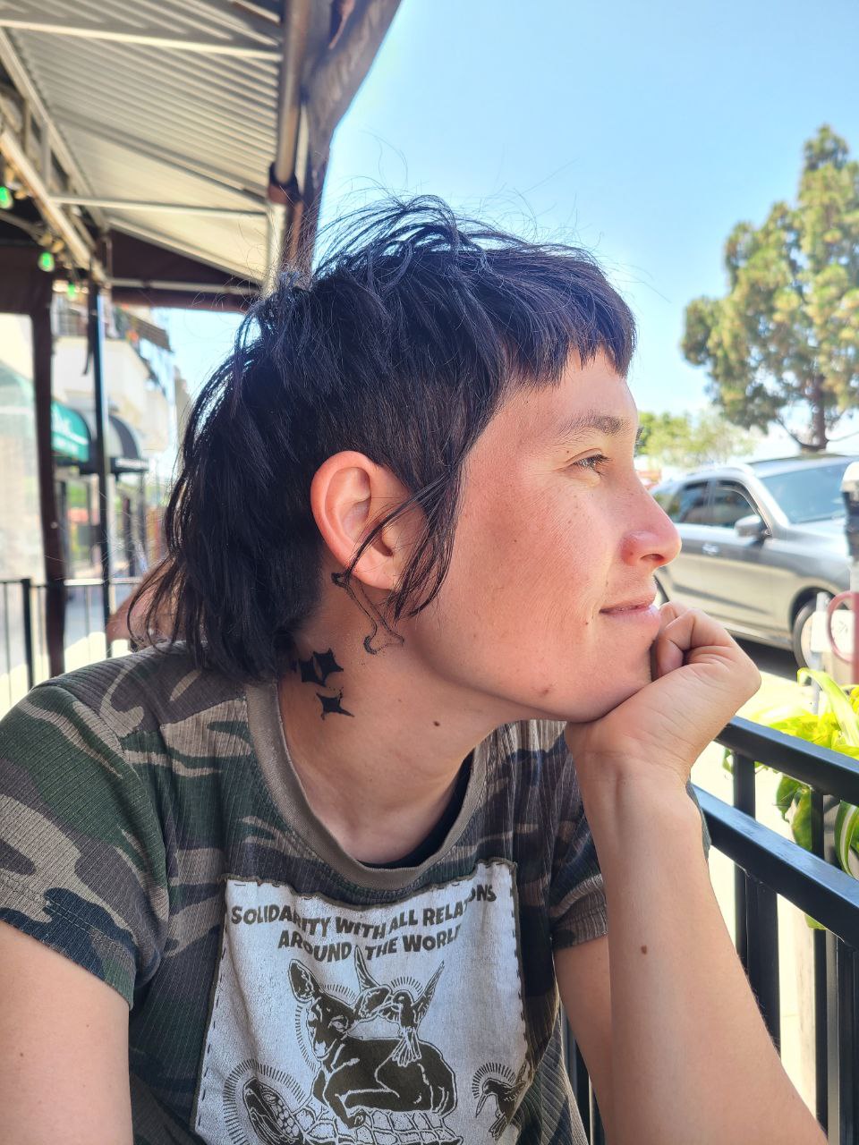 Amy (they) head turned towards the city with temporary ink tattoos on their neck of a bone and sparkles. Their shirt says, "Solidarity with all relations around the world" and has images of a turtle, a deer, and hummingbirds.