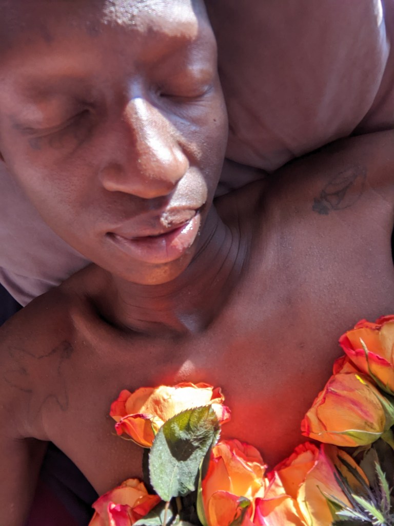 kuwa jasiri resting with eyes closed and roses places upon beauty's chest. Beauty's head rests on a pillow.