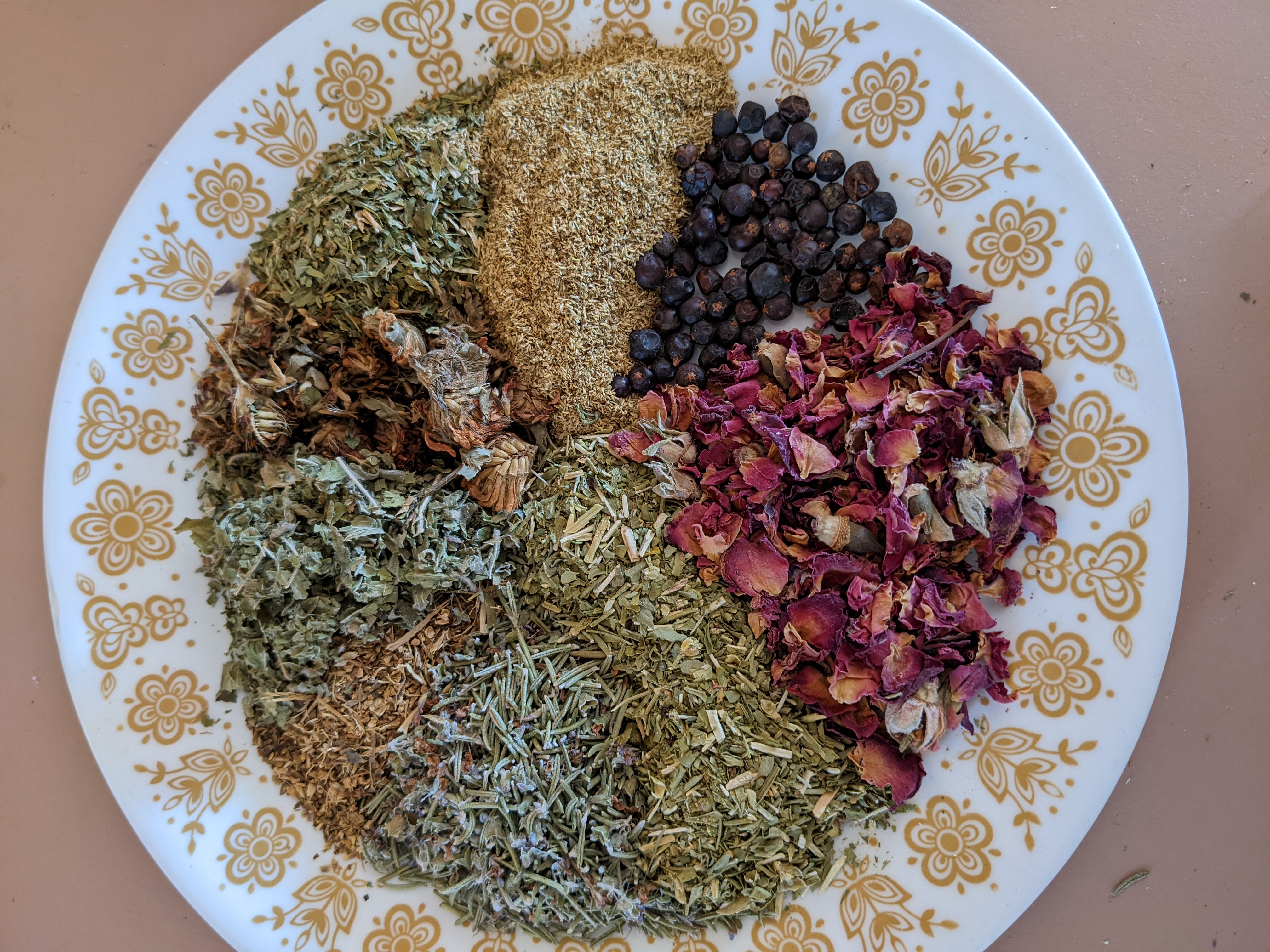 A plate of dried medicinal plants in the middle of the picture, with plant pieces, flower petals, and fruits. They rest on a white plate with yellow floral print around the rim. The plate rests on a pink surface.
