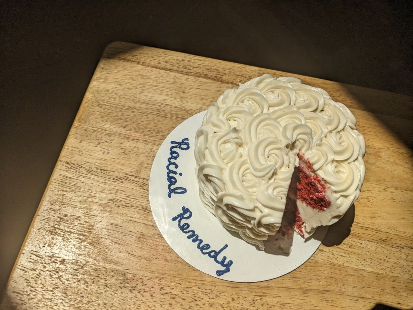 In the middle of the frame a white cake with swirled frosting and a slice cut out says below "racial remedy." It sits in a frame of sunlight on a table.
