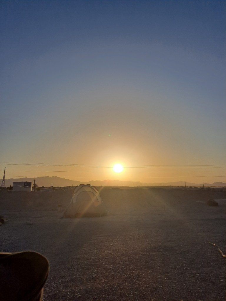 The Sun shines low in the horizon on a clear-skied day. There are mountains in the hazy distance. In the foreground, there is a tent set up amongst open land.