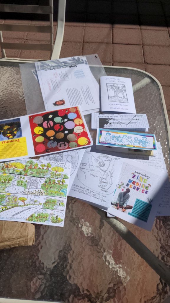 A collection of zines scattered over a glass table with a white metal frame. The glass is textured and slightly opaque. The ground is reddish brown tiles.