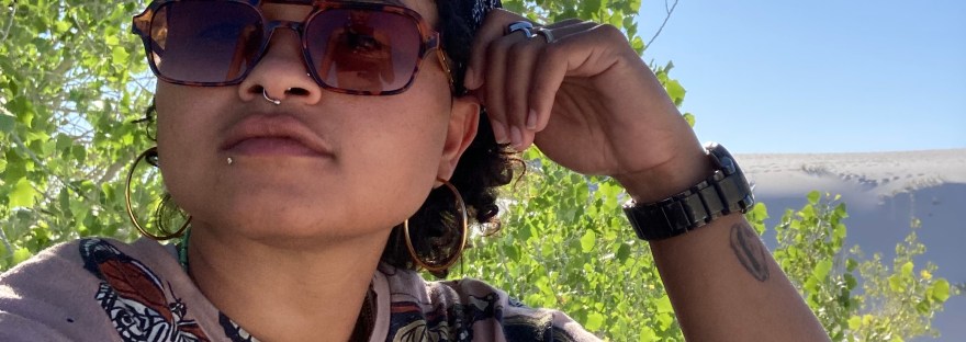 Taz (any pronouns) is sitting in white sands on a clear, blue skied day, in front of green bushes, with their head resting against their arm. Taz is wearing a butterfly printed shirt with a black bandana and brown sunglasses.